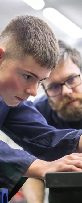 A lecturer and a student in navy-blue overalls working together in a mechanical engineering workshop. The lecturer is demonstrating how to use a tool on a metal component held in a vise. A lecturer and a student in navy-blue overalls working together in a mechanical engineering workshop. The lecturer is demonstrating how to use a tool on a metal component held in a vise.